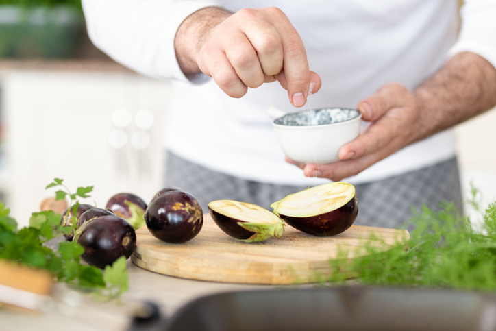 Kok bestrooid doorgesneden aubergine met zout, die op een houten plank liggen Daarnaasr hele aubergines en takjes groene kruiden 