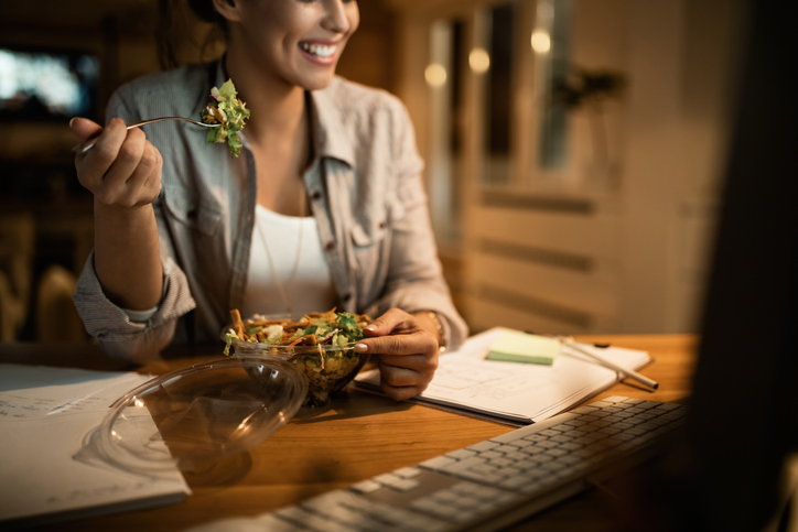 Vrouw aan tafel bij kunstlicht achter de computer met ee salade