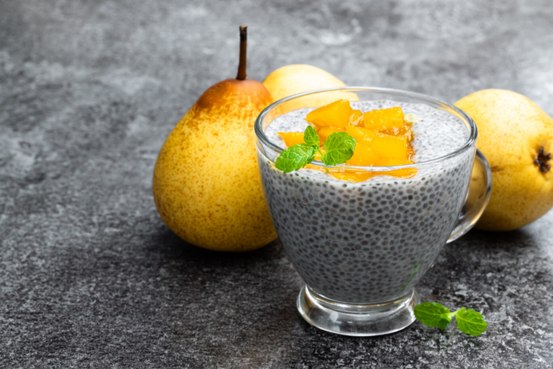 Glazen schaaltje met Chia-roompudding met stukjes stoofpeer in het glas en 2 ongeschilde peren ernaast 