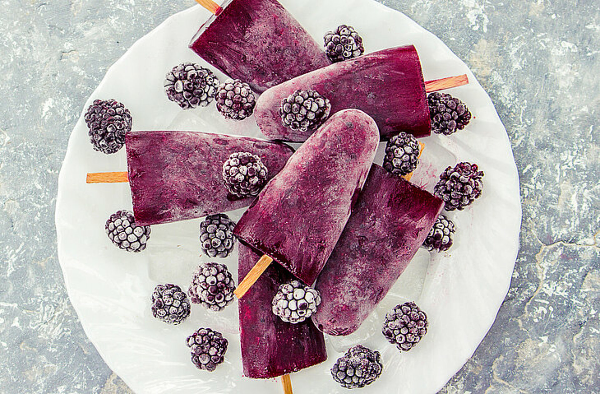 Wat kun je met grotere hoeveelheden rood fruit? Donkerrode ijsjes aan een srokje op een wit bord met bevroren bramen eromheen