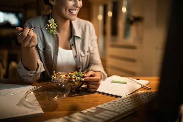 Vrouw aan tafel bij kunstlicht achter de computer met ee salade